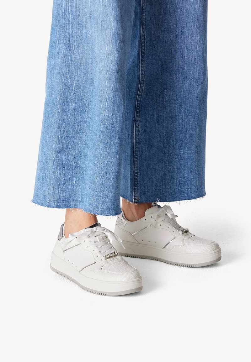 Zapatillas blancas con una superficie texturizada, punta redondeada y suela gruesa. Presentan cordones, herrajes decorativos y acentos grises. Llevadas con pantalones de mezclilla anchos.