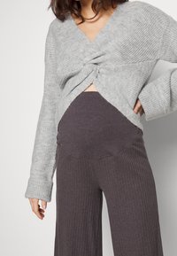 Femme portant un pull en maille torsadée gris et un pantalon large à taille haute gris foncé côtelé sur un fond uni.