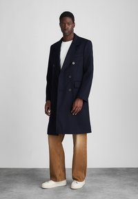 Tumed topeltkinnitatav mantel laiete kraedega, valmistatud villasegusest kangast. Omab suuri nuppe ja rinnaüksust, paari beige pükstega.