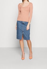 Rdzawy, prążkowany top z długim rękawem zestawiony z niebieską spódnicą z denimu o długości do połowy łydki, z centralnym rozcięciem. Modelka ma na sobie jasnopomarańczowe, szpiczaste szpilki.