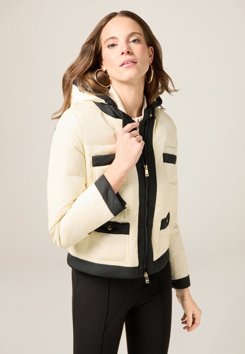 Padded jacket in cream and black, featuring a zip front, hood, and four front pockets with contrasting black details and trim.