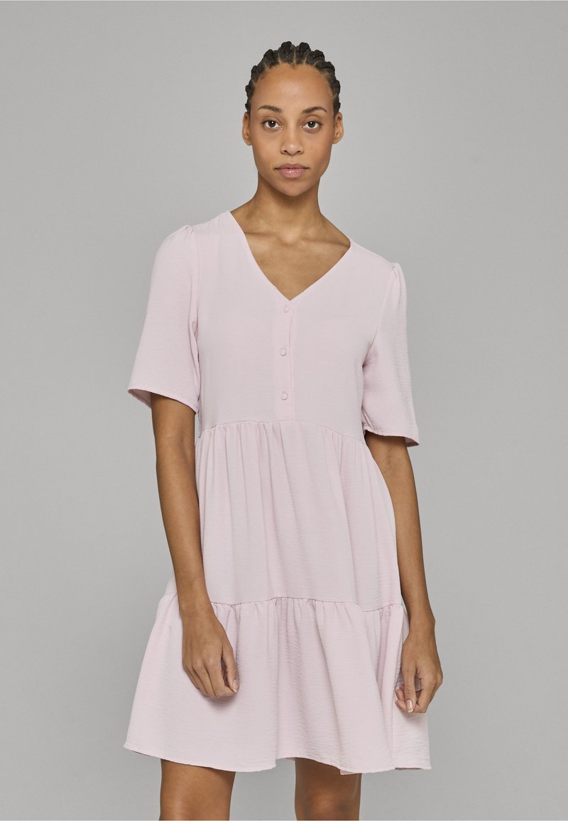 Donna con capelli intrecciati che indossa un vestito rosa chiaro a maniche corte con bottoni e gonna a balze, in piedi contro uno sfondo grigio uniforme.