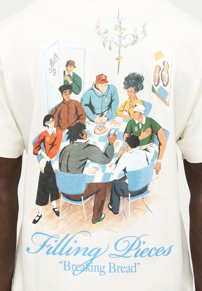 Επτά διαφορετικά άτομα μαζεμένα γύρω από ένα τραπέζι με ψωμί και ποτά, συνομιλώντας μέσα σε ένα δωμάτιο με πολυέλαιο και έργα τέχνης στους τοίχους.