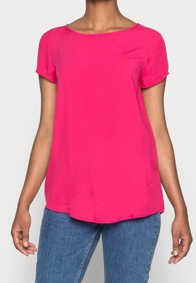 Haut à manches courtes en tissu rose vif, encolure ronde, texture douce, coupe décontractée, associé à un jean en denim, avec un ourlet arrondi.