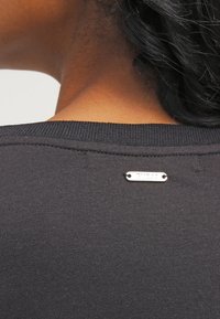 Camiseta de algodón negra con cuello acanalado y una pequeña etiqueta de marca plateada en la parte posterior, que muestra una textura suave y un diseño ajustado.