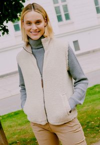 Gilet in pile morbido con colletto alto e tasche laterali, indossato su un maglione a collo alto grigio a coste, ambientato su uno sfondo verde all'aperto.