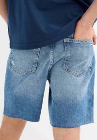 Shorts de denim en azul claro con un acabado desgastado, dobladillos deshilachados y dos bolsillos traseros. Combinados con una camisa azul marino, parcialmente visible.