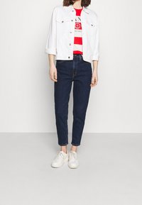 Valkoinen farkkutakki, punavalkoraidallinen t-paita tekstillä, siniset korkeavyötäröiset farkut ja valkoiset tennarit, kaikki esitetty neutraalia taustaa vasten.