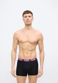 Άνδρας χωρίς πουκάμισο, που στέκεται στραμμένος προς τα εμπρός, φοράει μαύρα μποξεράκια Puma με ροζ ελαστική μέση με λογότυπο, σε λευκό απλό φόντο.