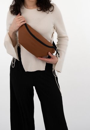 Femme en pull crème et pantalon noir tenant un sac bandoulière marron avec une fermeture éclair noire porté en travers du torse sur fond blanc.