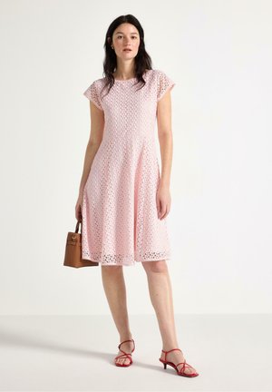 Kvinde iført en knælængde pink blondekjole, som holder en brun håndtaske og har røde, remmede sandaletter med hæl på, stående foran en ensfarvet baggrund.