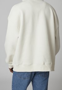 Sudadera color crema con un corte relajado, hombros redondeados y puños acanalados. Combinada con jeans de mezclilla azul. Textura suave, diseño minimalista.