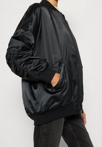 Blouson aviateur en satin noir avec poignets et taille élastiques, comprenant deux poches latérales et une fermeture éclair, texture lisse et épaules arrondies.