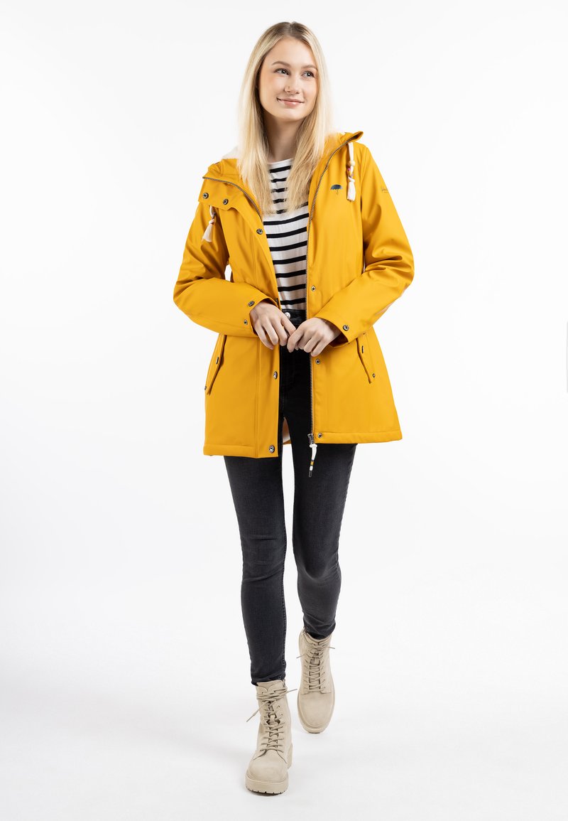 Giacca impermeabile gialla con cappuccio, dotata di tasche frontali e cordino di regolazione. Abbinata a una maglietta a righe, jeans scuri e stivali beige.