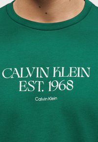 Πράσινο πουκάμισο με λευκό κείμενο "Calvin Klein EST. 1968" και μικρότερο "Calvin Klein" από κάτω, φορεμένο από άτομο που φαίνεται από τον λαιμό μέχρι το στήθος.
