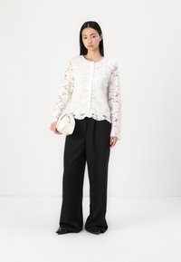 Blusa bianca in pizzo con maniche lunghe, scollo rotondo e orlo scallopato, abbinata a pantaloni neri a gamba larga e una piccola borsa bianca.