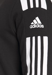 Felpa nera con design a tre righe bianche sulla spalla e logo Adidas. Materiale in cotone con una texture liscia.