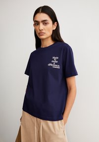 Frau mit langen dunklen Haaren, die ein navyblaues T-Shirt mit weißem Text "Made to make a difference" und beigen Hosen trägt, steht vor einer schlichten Wand.