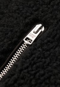 Zwart fleece stof met een grove textuur, met een zilveren rits langs één rand, gelabeld met "SUPERDRY" aan de ritstrekker.