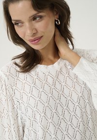 Blouse blanche texturée avec un motif en losanges, manches longues et col rond. Le modèle met en valeur un tissu doux et fluide.