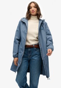 Jasnoniebieska wodoodporna kurtka z kapturem, suwakiem z przodu i bocznymi kieszeniami. Posiada teksturowane logo na klatce piersiowej oraz kontrastowe szwy.