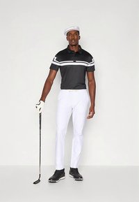 Svart pikétröja med vita horisontella ränder, vita byxor, svarta golfskor och en vit keps. Håller i en golfklubba i en neutral pose.