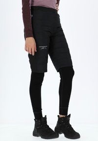 Black padded shorts with logo, set against black thermal leggings. Black hiking boots feature laces and textured sides, standing on a light background.