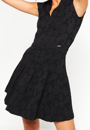 Kobieta ubrana w czarną, kwiatową, teksturowaną sukienkę bez rękawów z dopasowaną talią i rozkloszowanym dołem, pozująca z uniesionymi ramionami.