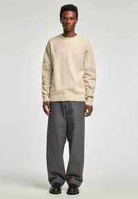 Bēša adīta džemperis ar apaļu apkakli, kopā ar pelēkām svītrainām platām biksēm un melniem apaviem, stāvot pret vienkāršu fonu.