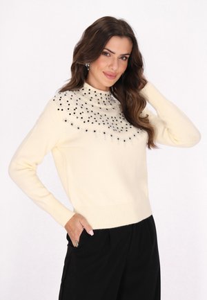 Woman with wavy brown hair wearing a cream sweater with black bead embellishments and black pants, posing with one hand in pocket and the other on head.