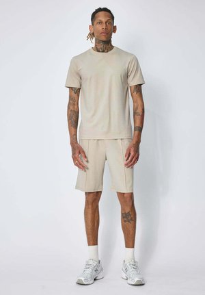 Homme avec des tatouages portant un t-shirt beige et un short, des chaussettes blanches et des baskets, debout devant un fond clair uni.