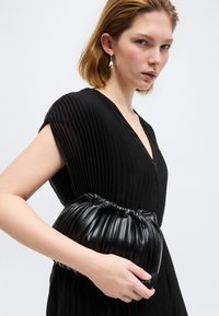 Νεαρή γυναίκα με κοντά μαλλιά, ντυμένη με μαύρο πλισέ φόρεμα και καρφωτά σκουλαρίκια με μαργαριτάρια, κρατώντας μια μαύρη πλισέ δερμάτινη τσάντα φάκελο.