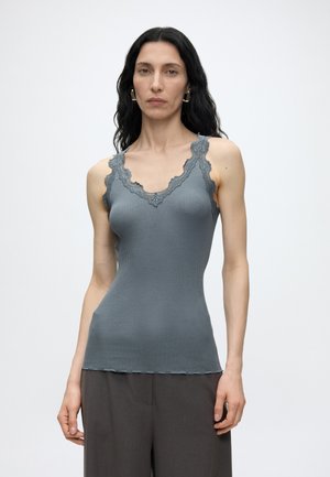 Femme aux longs cheveux foncés portant un haut gris côtelé sans manches avec un bord en dentelle et un pantalon large foncé, debout devant un fond blanc uni.