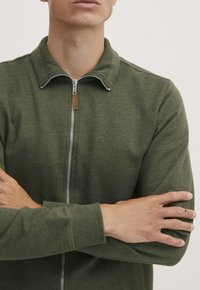 Grønn zip-up jakke laget av mykt stoff med krage, med en sølv glidelås og et treskilt. Ribbestrikkede mansjetter.