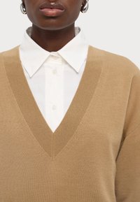 Pull en maille beige à col en V superposé sur une chemise blanche à col. Tissu texturé et coupe décontractée. Boucles d'oreilles argentées en forme d'anneaux visibles.