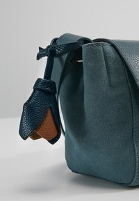 Teal läderaxelväska med texturerad yta, med en distinkt tofs och en kontrasterande brun accent på botten.