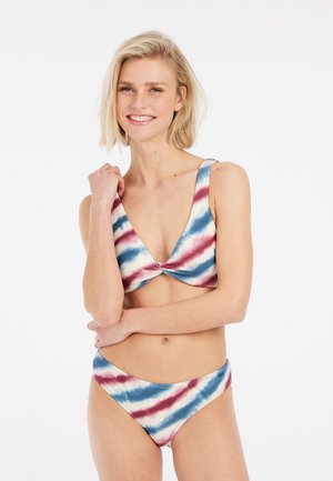 Šypsosi moteris su trumpais šviesiais plaukais, vilkinti mėlyną, raudoną ir baltą tie-dye bikinį, stovinti sukryžiuotomis rankomis prieš baltą foną.