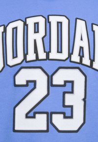 Šviesiai mėlynas džersis, kuriame yra didelis baltas tekstas "JORDAN" su juodu kontūru ir skaičius "23" baltos spalvos, abu su tekstūruotu siuvinėjimu.