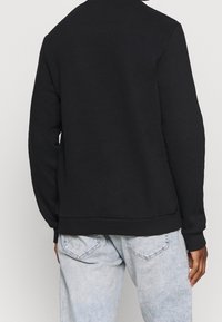 Sweatshirt noir avec encolure ronde et ourlet côtelé. Confectionné en tissu doux, il présente une coupe décontractée et des manches longues. Fond neutre.