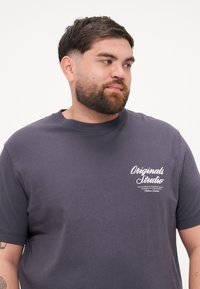 Námořnická modrá bavlněná tričko s krátkým rukávem, kulatým výstřihem a bílým nápisem "Originals Studio" na levé oblasti hrudi. Hladká textura.