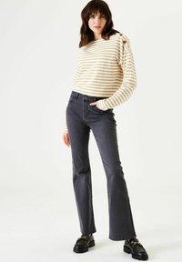 Gestreiftes Creme- und Gold-Longsleeve mit Schulterknöpfen, kombiniert mit dunkelgrauen Schlagjeans und schwarzen Mokassins mit einem goldenen Akzent.