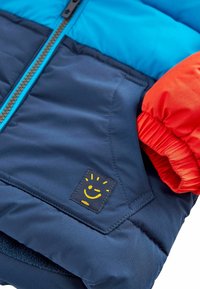 Giacca imbottita con sezioni blu e navy, polsini elastici arancioni e un logo con faccina sorridente gialla su una toppa scura. Dettaglio con cerniera visibile.