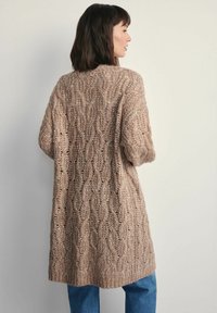 Sweter kardigan w jasnobrązowym kolorze, z wzorem w warkocz i ażurowym wykończeniem. Luźny krój z krótkimi rękawami, sięgający do połowy uda.