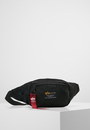 Czarna nerka Alpha Industries z kieszeniami na zamek i czerwoną metką z logo, położona na białej powierzchni na szarym tle.