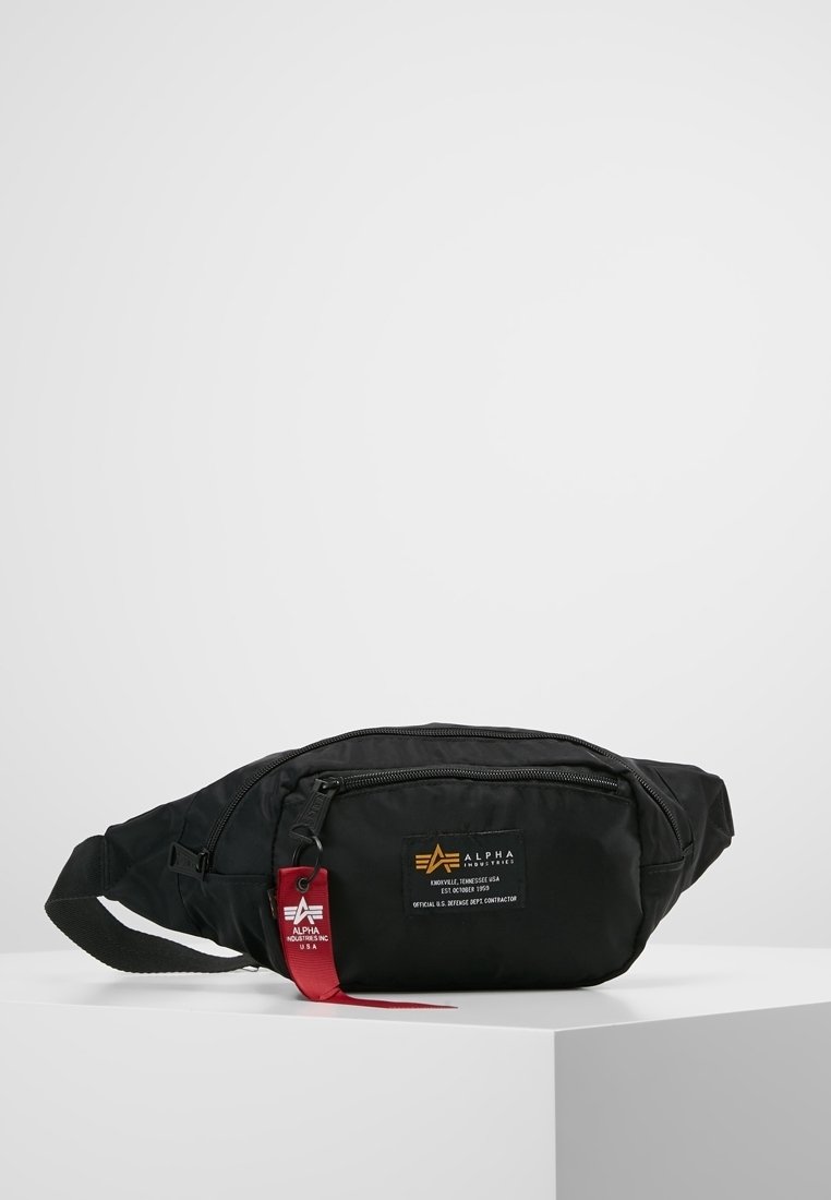Sac banane noir Alpha Industries avec poches zippées et étiquette de tissu rouge à la marque, posé sur une surface blanche devant un fond gris.