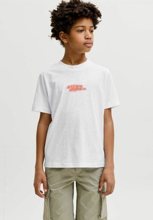 Jongen met krullend haar, gekleed in een wit Jack & Jones T-shirt en beige cargoshort, staand tegen een effen lichte achtergrond.