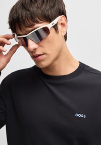 Akiniai nuo saulės su baltais rėmeliais ir rudais lęšiais. Dizainas apima sportinę formą ir prekės ženklo pavadinimą "BOSS" šone.