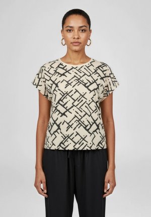 Kvinde med kort hår iført guld øreringe, en beige top med sorte abstrakte linjer og sorte bukser mod en ensfarvet baggrund.