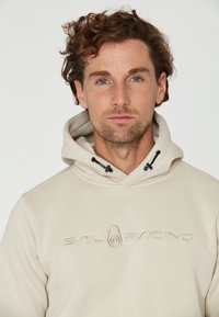 Sudadera beige con un logo texturizado "SAIL RACING" en el pecho, con capucha ajustable y fabricada en suave algodón.