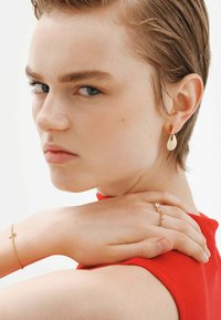 Pendientes de gota de oro, pulsera de cadena delicada con una cruz y anillo con gemas, exhibidos sobre una blusa roja y un fondo neutro.